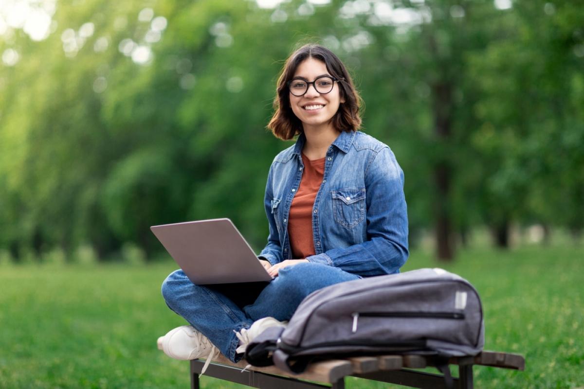 Female Student Sitting On Bench Outdoors With Laptop